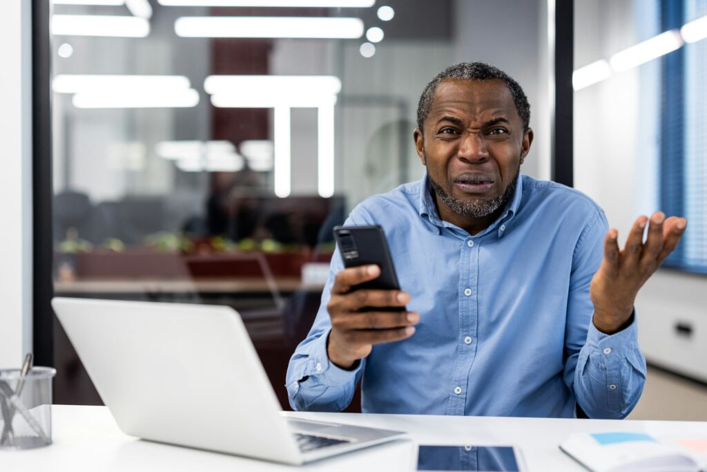 Mature businessman showing frustration at workplace using smartphone inside office